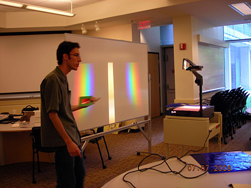 The teacher stands next to a board where rainbow images are visible.