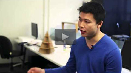 Instructor William Li sits in front of a long white table. Hes gesturing with one arm. There are two computer monitors on the table, along with a three dimensional object constructed out of cardboard.