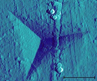 A sharp probe has left a triangular indentation in a surface, shown using scanning electron microscopy.