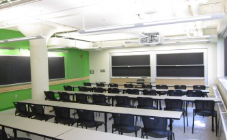 Four rows of grey tables with black chairs. Sliding blackboards at the front and along the left hand side of the room. The wall to the left is painted green.