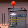 A tuning fork sits next to a rectangular tank of water with a clear bottle in it. A red sphere on a stick rests on top of the bottle.