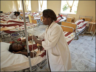 Woman standing over another lying in a hospital bed next to an IV drip.