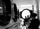 Photograph of people doing laundry. A lady explains that the lack of space in the city creates the unfriendly atmosphere inside the laundromat.