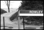 Photo of a sign labeled 'Rowley' on a train station platform with a chain-link fence running behind it.