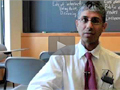 Dr. Mahajan sits in a student's desk in a classroom with a chalkboard behind him.