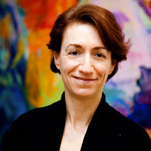 A photo of a smiling woman standing in front of a very colorful wall painting.